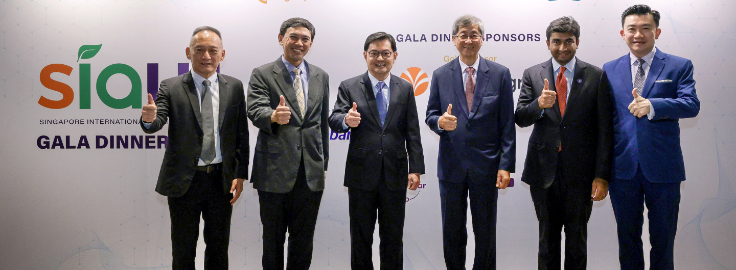 Six people in suits give thumbs up in front of a "SIA Singapore International Gala Dinner" backdrop.